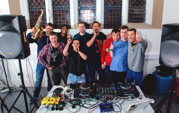 Full group of 7 musicians behind the decks with Luke and Ben from Drake Music, celebrating and giving thumbs up