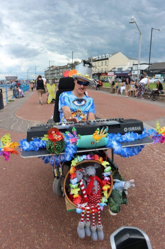 Steve is pictured in his wheelchair on the seafront. It is adapted with a large keyboard and bass drum and is decorated with colourful flowers