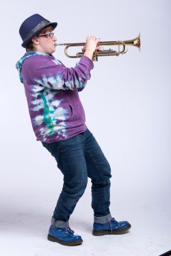 Robyn is shown in profile playing a trumpet, leaning back, knees bent, wearing a purple hoodie, jeans and trilby hat