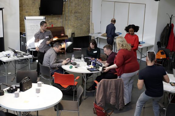 Six people are getting involved with making music using technology. They loosely gathered around a table, but are each doing something different