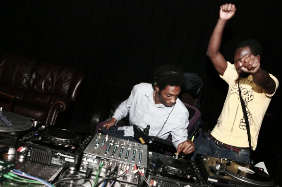 A young black man DJ's with decks and another man dances alongside him