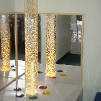 Image: Corner of room with mirrored walls and tall clear plastic tube containing bubbles lit up from below.