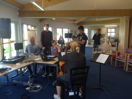 Three musicians playing together on the cello, iPad and Mixing Desks, while composers stand nearby, listening