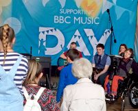 A view from behind the crowd watching a groups of young musicians perform in front of a big BBC Music Day banner
