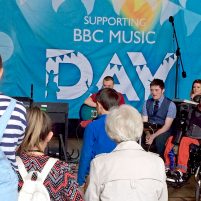 A view from behind the crowd watching a groups of young musicians perform in front of a big BBC Music Day banner