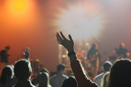 Image of a concert from the viewpoint of the crowd, with waving hands, bright lights & musicians blurred in the distance