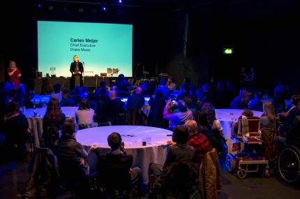 Carien Meijer introduces Drake Music's work onstage, in the foreground are tables full of people