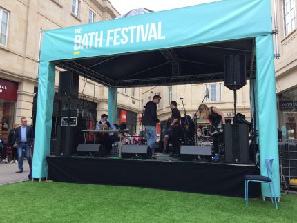 A blue marquee houses a small stage where the performers are gathered with their instruments