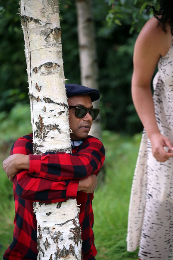 A man wearing sunglasses and a checked shirt hugs a tree tightly.