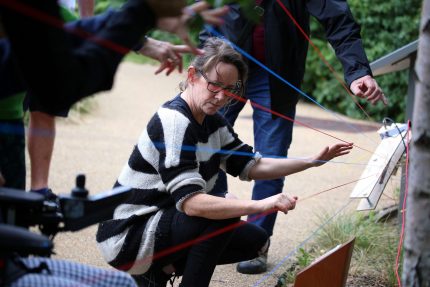 A group of people crouch next to multi-coloured strands of wool running between trees.