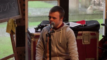 A young man sits onstage, singing into a microphone wearing a grey hoodie