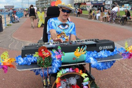 Steve is pictured in his wheelchair on the seafront. It is adapted with a large keyboard and bass drum and is decorated with colourful flowers