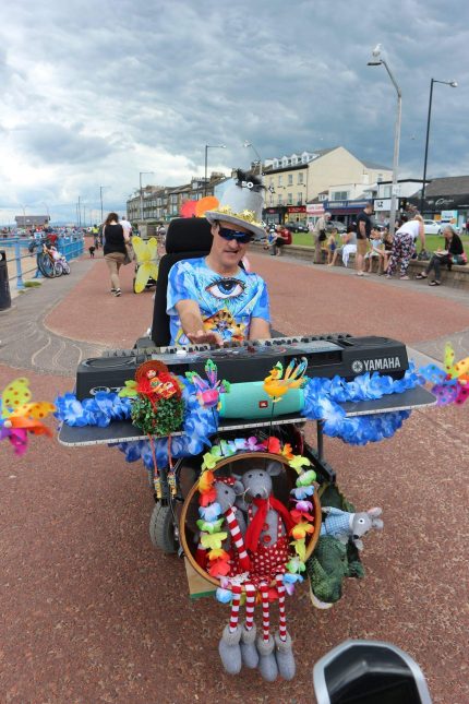 Steve is pictured in his wheelchair on the seafront. It is adapted with a large keyboard and bass drum and is decorated with colourful flowers