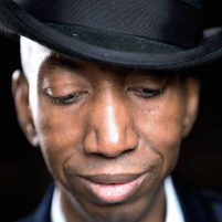 Dike wears a black trilby hat and his black suit and white shirt are just visible. He looks down with the brim of his hat pulled low and a slight smile