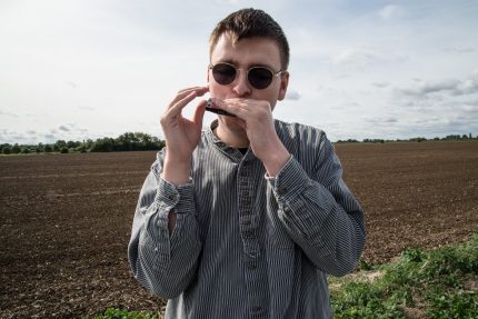 Oliver stands on the Fens before a ploughed field. He is wearing round shades and looking into the camera as he plays his harmonica