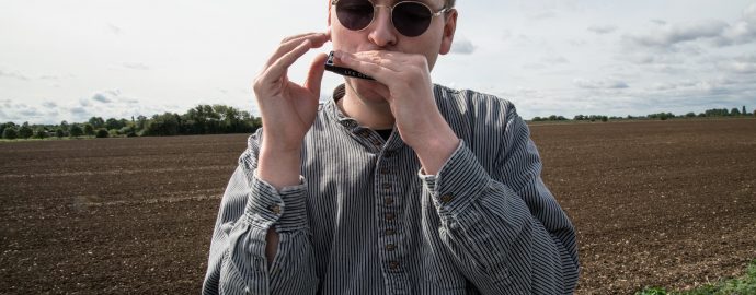 Oliver stands on the Fens before a ploughed field. He is wearing round shades and looking into the camera as he plays his harmonica