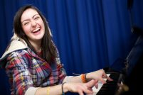 Sarah sits at the piano with a big smile, she has long brown hair and wears a plaid shirt.