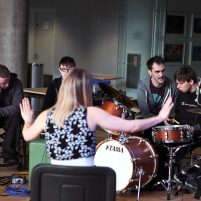 A music leader holds out both arms with her palms up as she tells the group of young musicians to stop playing