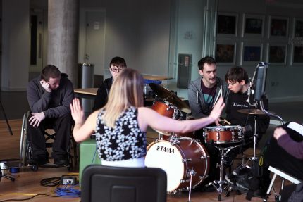 A music leader holds out both arms with her palms up as she tells the group of young musicians to stop playing