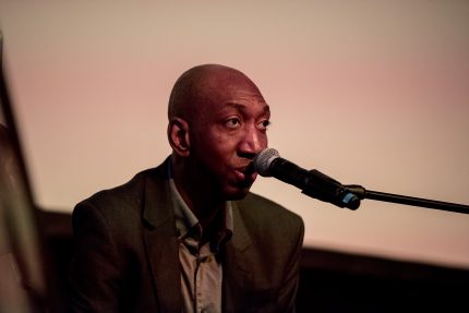 Dike sings into a microphone wearing a dark blazer