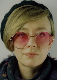Sally looks directly at the camera, her lips pressed together, not smiling. She has a sweeping blond fringe and hexagonal pink tinted glasses.