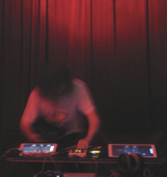 An atmospheric dark, blurred photograph shows Robbie in action making music using technology, performing in front of a red velvet curtain