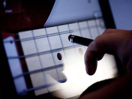 A hand holding an electric pen hovers over an ipad with a guitar fret visible on the screen