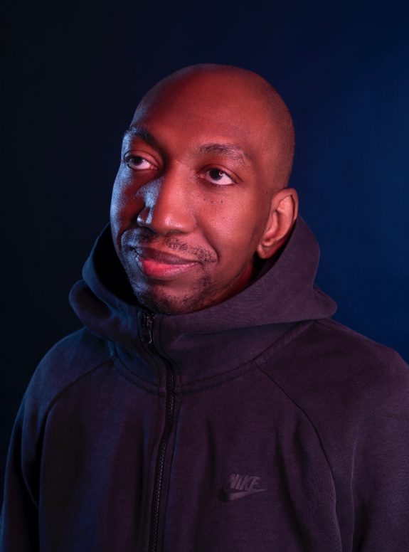 Dike wears a Nike hoodie and looks off camera with a soft, slightly smiling expression. Shot inn low light against a dark backdrop, the image has a rich tone.