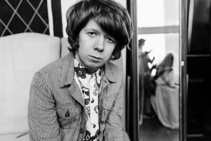 A black and white photo of James wearing stylish sixties clothing. He has a mop top hair cut and looks at us quizzically.