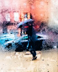 A person is seen through a rainy window, walking down the street holding an umbrella
