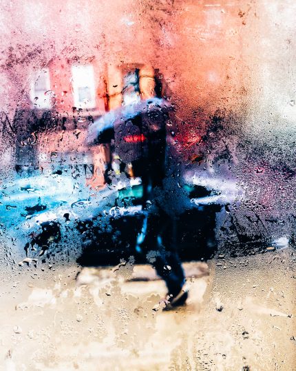A person is seen through a rainy window, walking down the street holding an umbrella