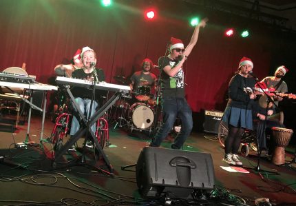 A band onstage wearing christmas hats and the lead singer has his hand in the air