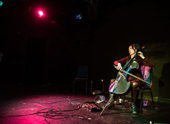 Jo-anne plays cello on a stage lit in pink