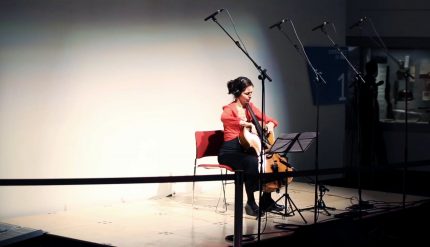 A cellist sits alone on a wide stage, lit brightly