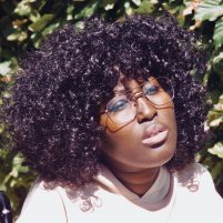 Miss Jacqui is a young black woman with glasses. She has spiral curls just reaching her shoulder, a very pale yellow top and a thoughtful and almost dreamy expression.