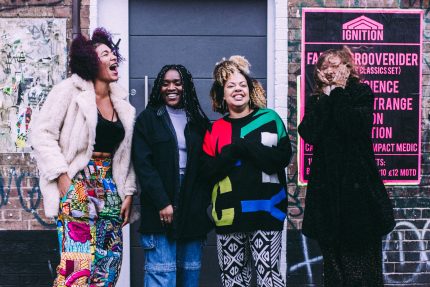 Four women of colour stand together in front of live music posters. They are dressed in colour, pattern and black and are smiling and joking.