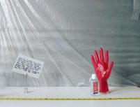 A red rubber glove stands up on a plain surface, with antibacterial gel on and next to it. This looks like a strange laboratory of some kind with plastic sheeting and measuring implements around.