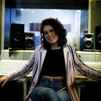 Joy smiles as she leans back against a mixing desk in a studio. She is a young white woman with curly dark hair.