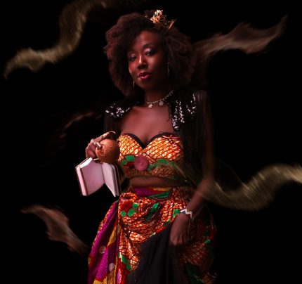 Radikal Queen is a young black woman wearing a skirt and top made from brightly-coloured traditional African wax print fabric. She has an afro and wears a small crown and a slight smile.