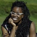 Ruth is a Black woman with long hair and white framed glasses. She smiles and rests her head on her hand.