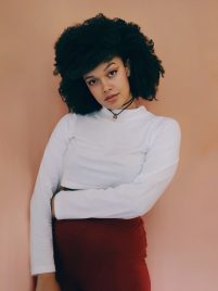 Elle is a young Black woman wearing a white long scleeve top. She has one arm folded across her waist and her head is tilted, eyebrow raised.