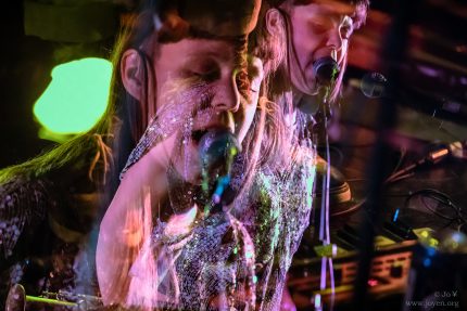 A double exposure of R.Dyer singing. She has long brown hair and a blunt fringe. Her eyes are closed and she wears a sequin top.