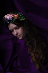 Elinor is wrapped in a rich purple velvet cloth, she has her eyes closed and her head turned to the side, pressing her cheek to the fabric. She is a white woman wearing a crown of flowers with long brown hair.