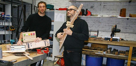 Two white men in a workshop environment are striking funny poses to show off unusual instruments they have built