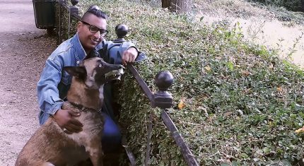 An asian man wearing a denim jacket, smiling as he crouches down and strokes a large dog