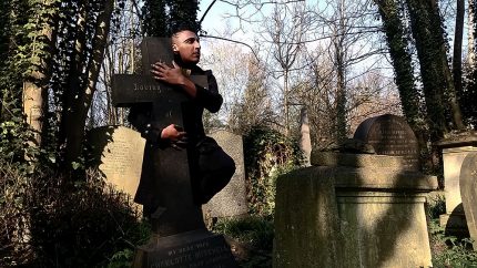 Patrick stands in a graveyard lit by sunlight. He wears a smart coat and has black hair