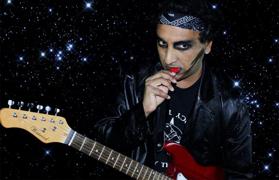 Musician Patrick is wearing a bandana and black eyeliner, holding an electric guitar and pick.