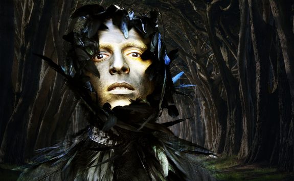 a man's face, in gold paint, he is in a fantastical sort of scene, with black feathers around his face and dark trees behind him.