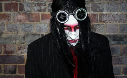 A man with black hair, his face painted white with a red stripe across the eyes and black liner. He is wearing dark goggles on his head and looking up at the camera with a hint of menace.