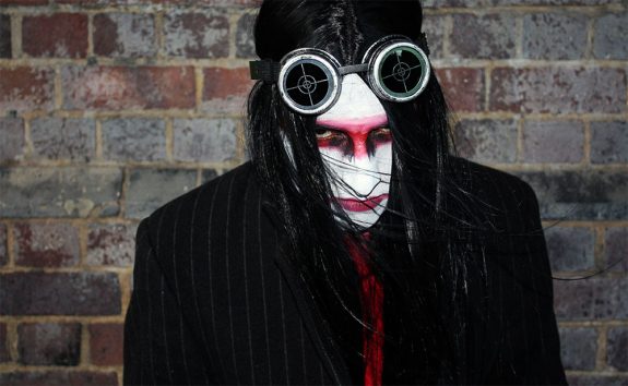 A man with black hair, his face painted white with a red stripe across the eyes and black liner. He is wearing dark goggles on his head and looking up at the camera with a hint of menace.